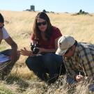 Woman talking to two men. All three are squatting in a field of low, dry grass, with farm buildings at the horizon.