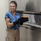 Woman at a large commercial oven, smiling and holding a loaf of freshly baked bread. It looks yummy! 