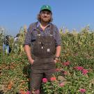 A young man wearing overalls and a ball cap stands among tall bushes bearing pink, orange and wine-colored flowers