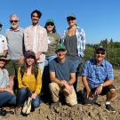 10 men and women in farmwork clothes pose in a field, with a blue sky behind them.