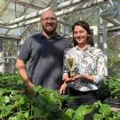 A woman holds a small gold-colored trophy. She stands with a man in a greenhouse. In front of them are trays with hundreds of 4-inch pots with small green plants growing