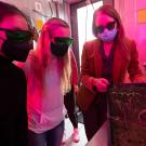 Three women wearing goggles inside a small room with reddish-purple light look at plant sprouts growing in a tray.