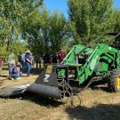 Large green tractor-like machine with an arm extended to the right side, at the edge of a row of green-leafed trees in an orchard. People standing on either side, watching.