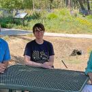 A man and two women sitting at a metal table with a garden in the background